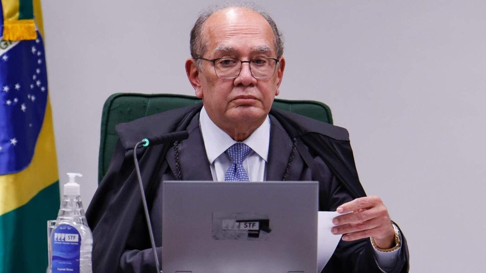 Gilmar Mendes, ministro do STF, de toga, sentado à mesa do plenário com notebook e bandeira do Brasil ao fundo