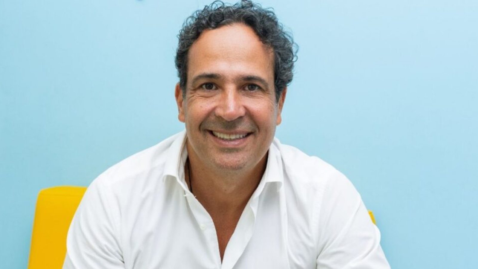 Pablo Teixeira, CEO da +Pet, de camisa branca, sorrindo contra fundo azul e cadeira amarela