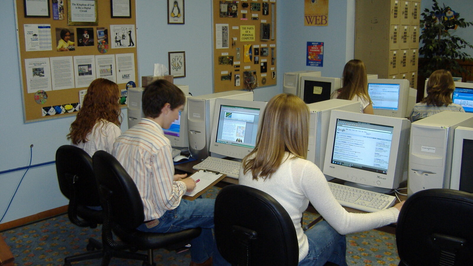 Estudantes trabalhando em laboratório de informática com monitores, em sala de aula iluminada.